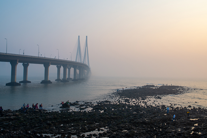 A long-distance photo of the Bandra-Worli Sea Link bridge in Mumbai, India, shrouded in a hazy or misty atmosphere, likely at sunrise or sunset.
