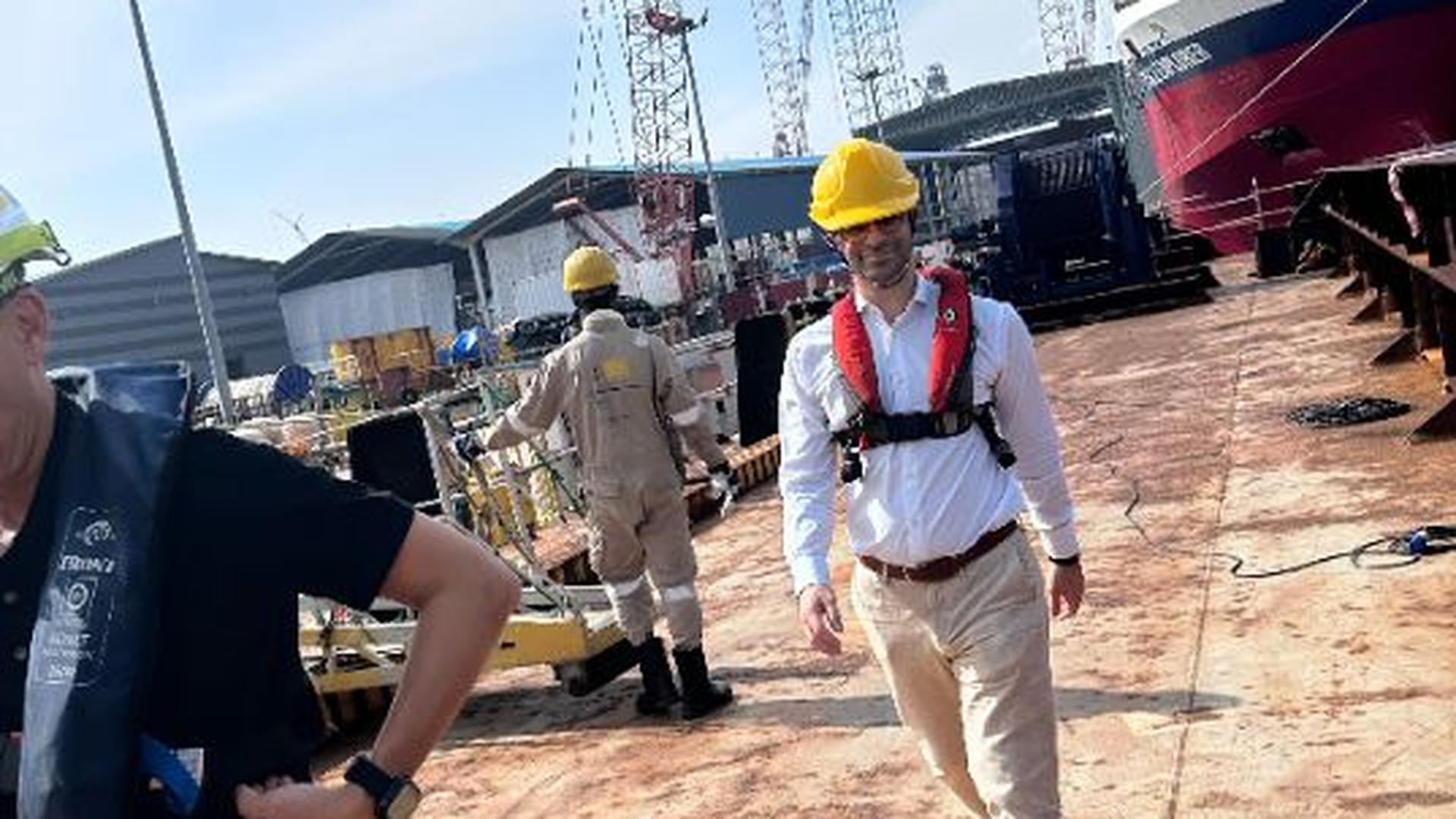 Three men in a shipyard or dock setting. The man in the foreground is walking towards the camera, wearing a white shirt, light tan trousers, a yellow hard hat, and an orange and white life vest. Another worker in a tan jumpsuit and yellow hard hat is walking away in the middle ground, and a third person is partially visible on the left, also wearing a life vest.