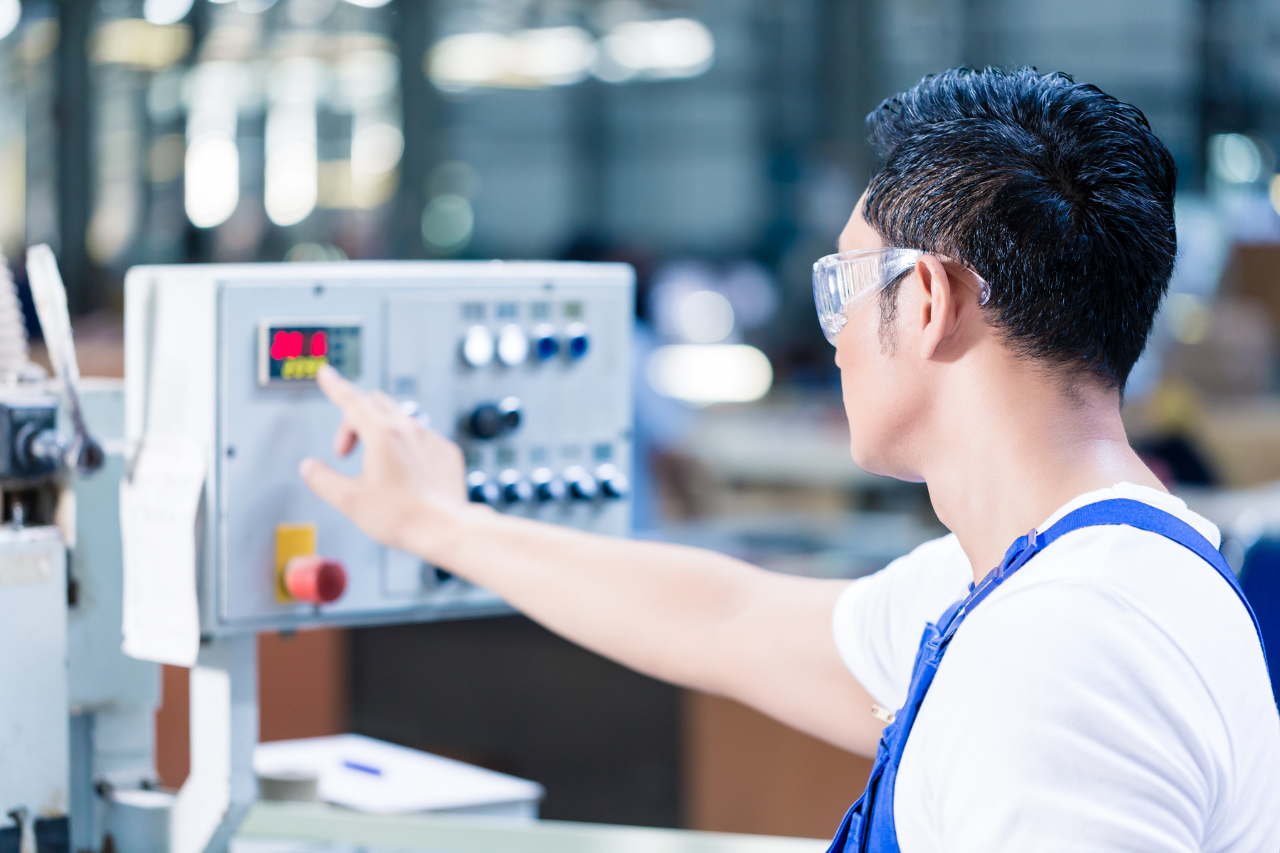 A worker wearing safety glasses and a white shirt with blue overalls operates a machine control panel in a factory or workshop, pointing to a digital display.