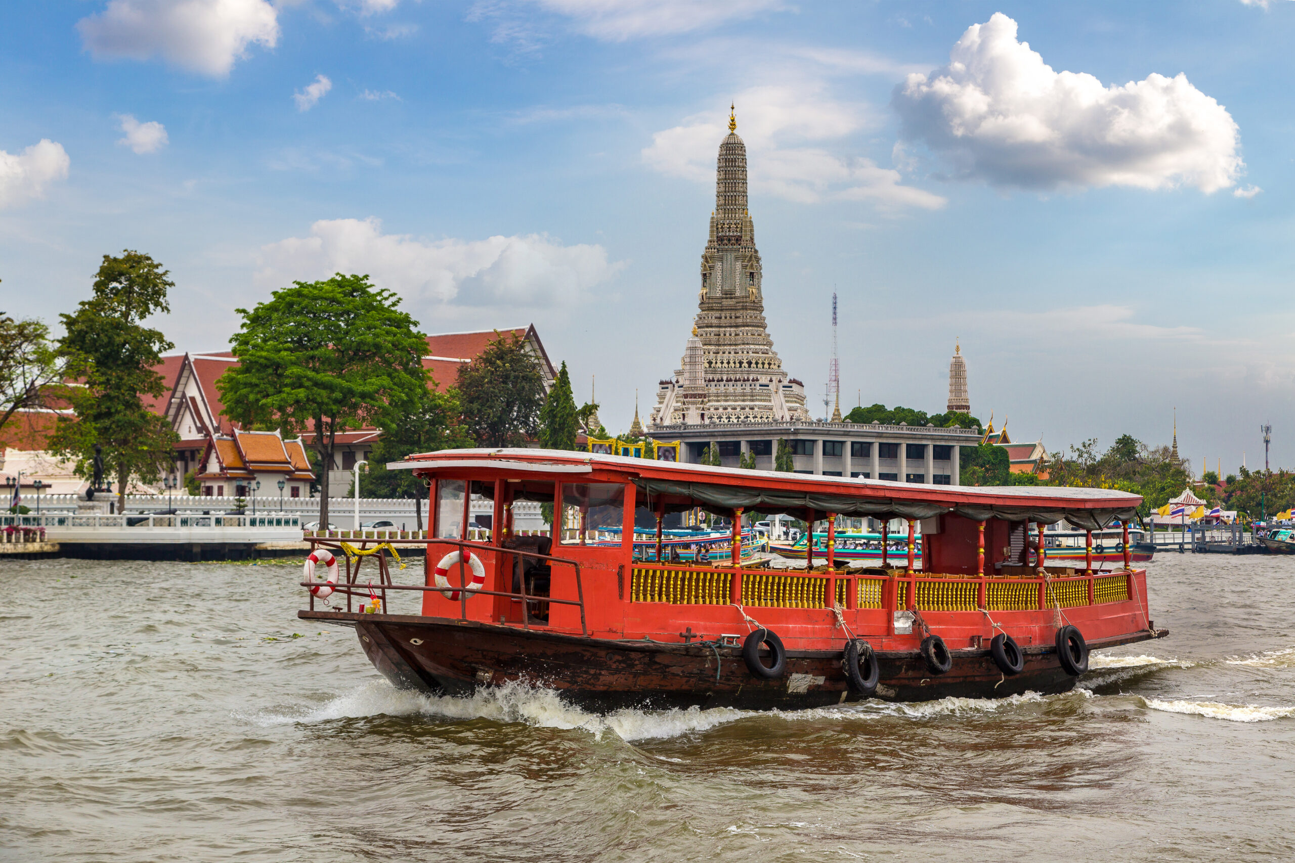 A red passenger boat cruises along the Chao Phraya River in Bangkok, Thailand, with the ornate spire of Wat Arun temple rising in the background under a partly cloudy sky.